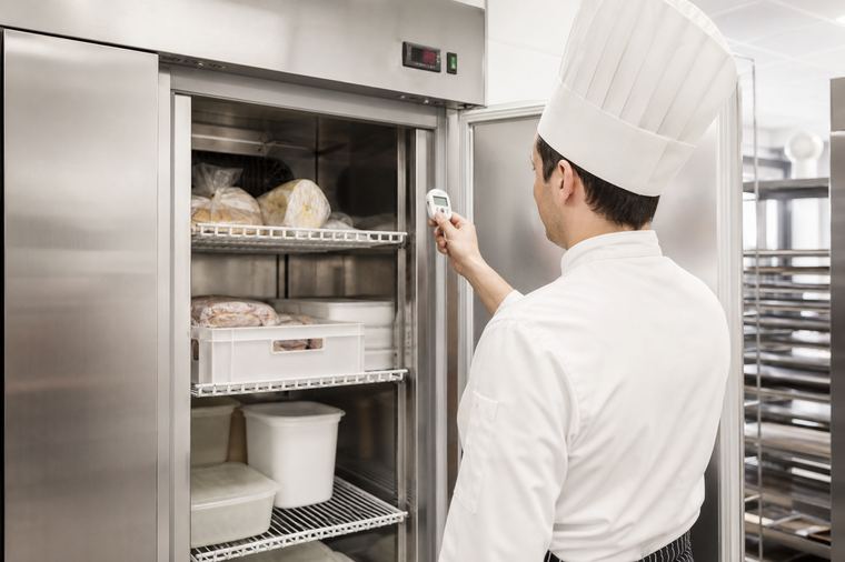 Sala di un locale con tavoli, sedie e cucina aperta sullo sfondo chiaro.